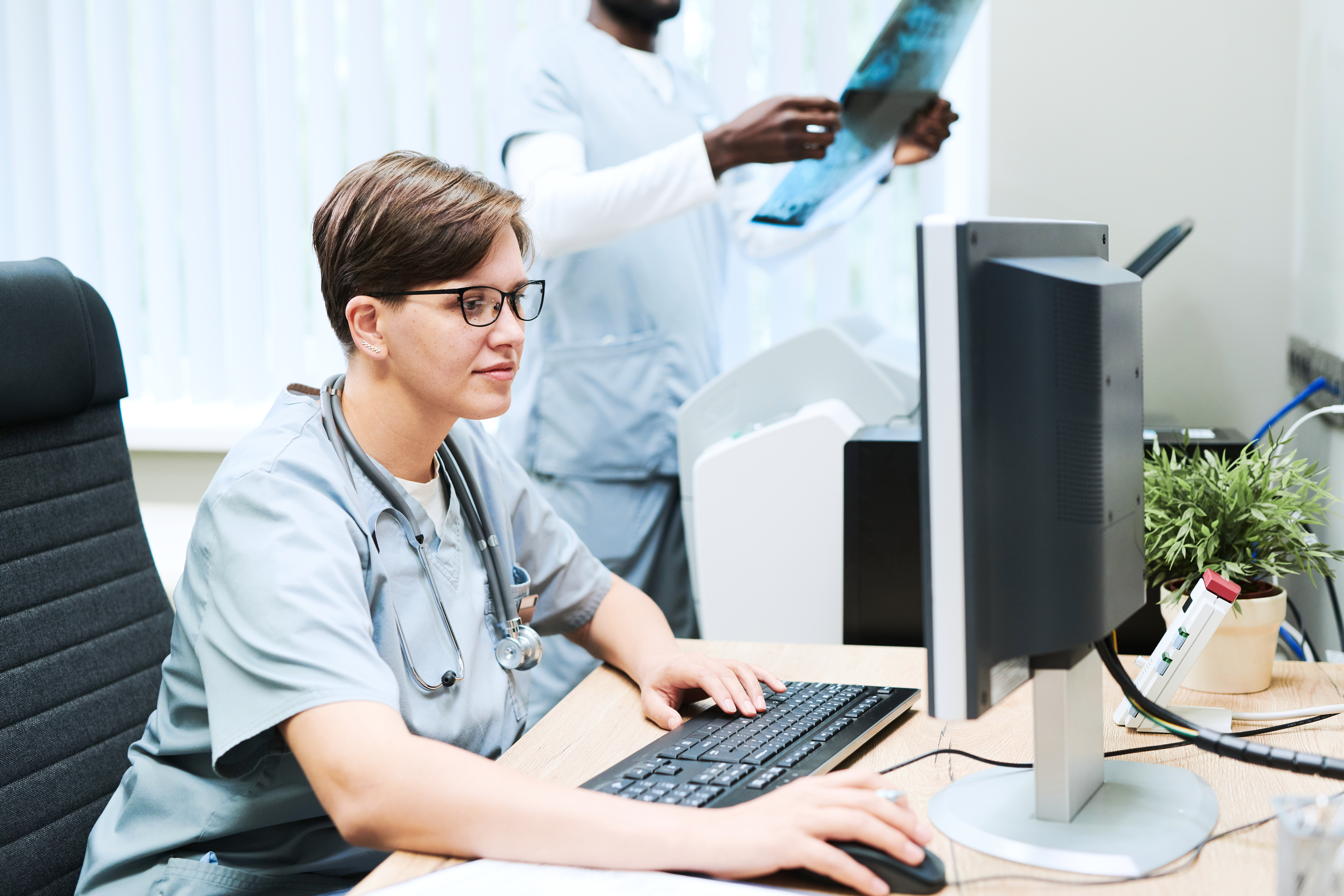 Doctor working with computer in office Content female doctor with stethoscope around neck working with computer in office while her colleague analyzing x-ray image