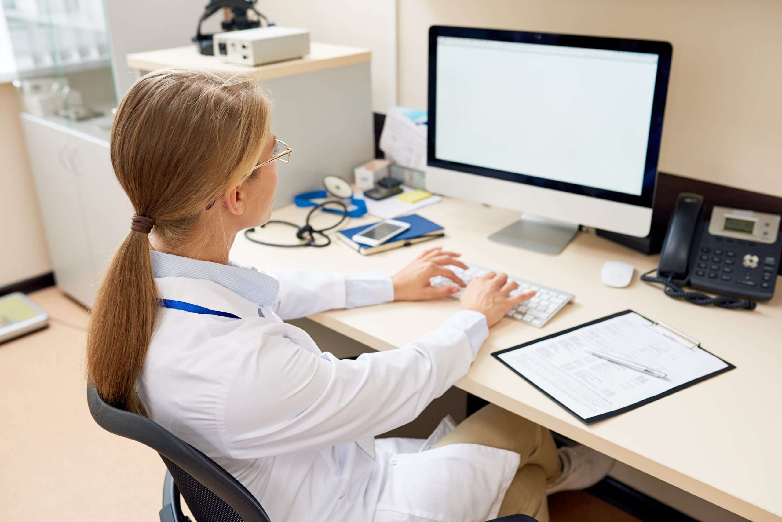 Female Doctor Working at Desk Back view portrait of blonde female doctor sitting at desk in office working with computer, typing and looking at blank white screen, copy space