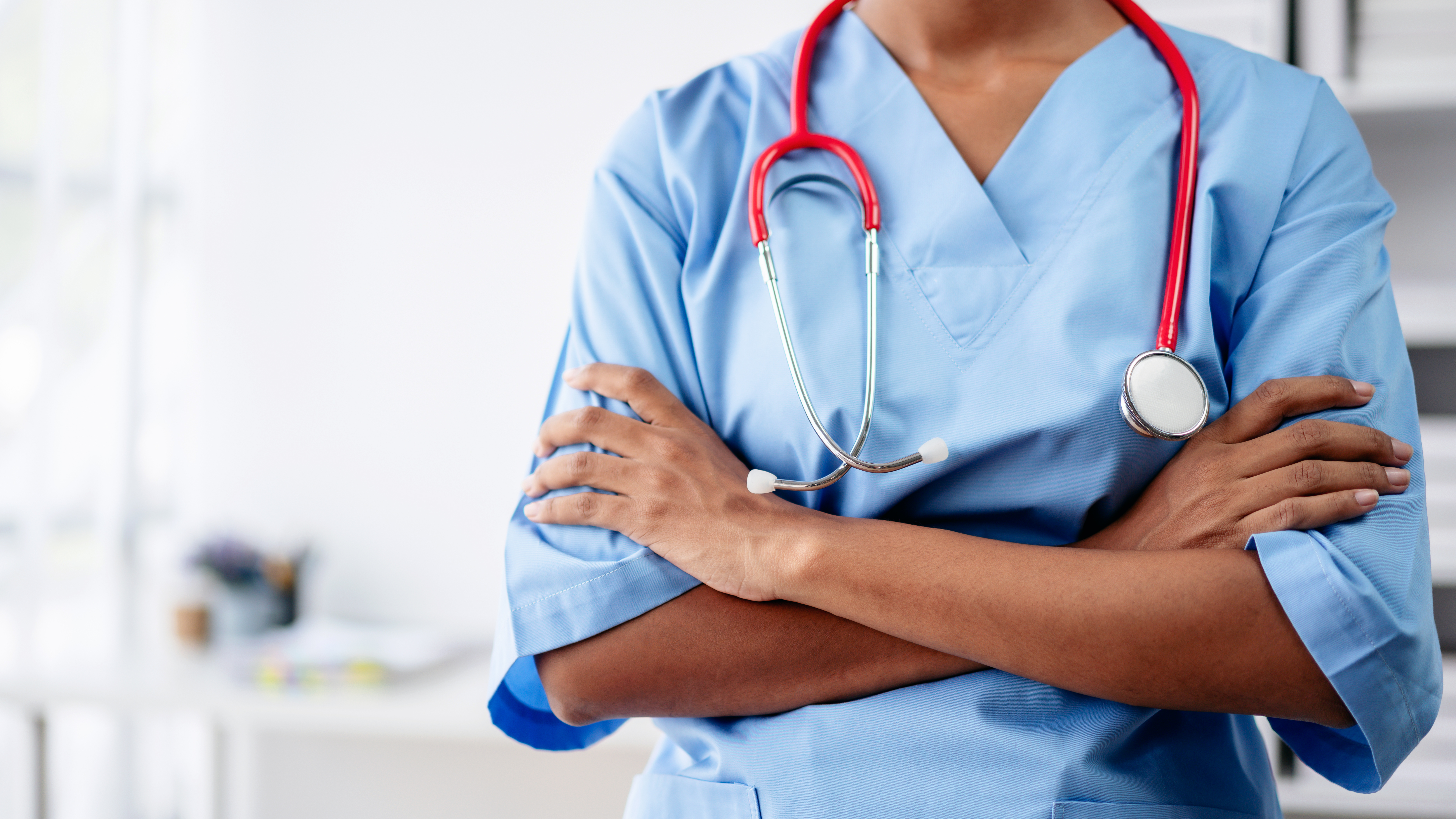 A woman in a blue scrubs shirt is holding a stethoscope and has her arms crossed
